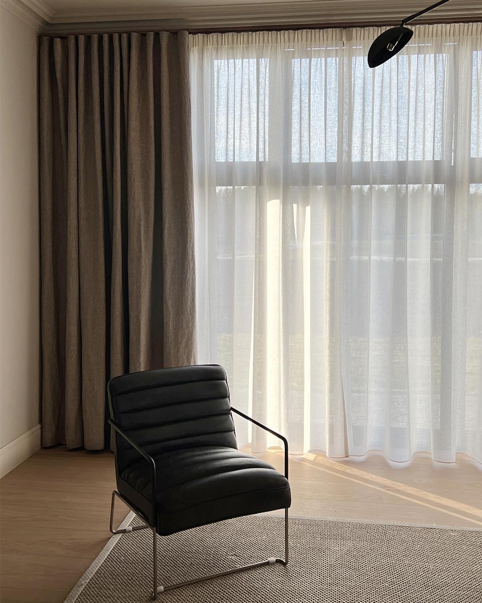 Lys stue med dobbel gardinskinne og to lag med gardiner. En sort skinnstol i forgrunnen, Foto.